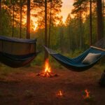 Camping hammocks set up between trees at dusk with bug netting, pillows, and a glowing campfire in the forest.