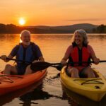 Senior couple kayaking on a peaceful lake in sit-on-top kayaks wearing life jackets, with scenic sunset and forest backdrop.