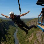 Bungee jumper mid-fall above a canyon with visible safety harness and launch crew on platform.